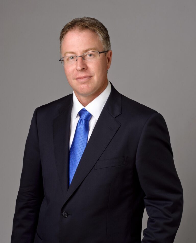 Man in a dark suit and blue tie, an executive professional, stands against a plain grey background.