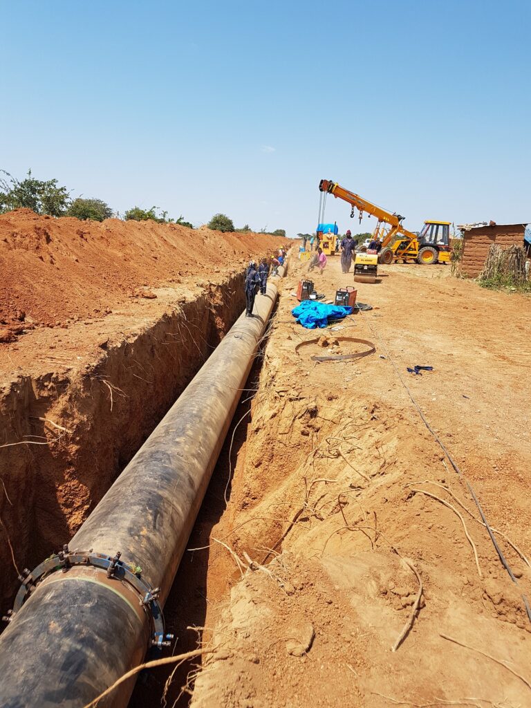 Construction workers install a large pipeline in a trench with equipment under a blue sky.