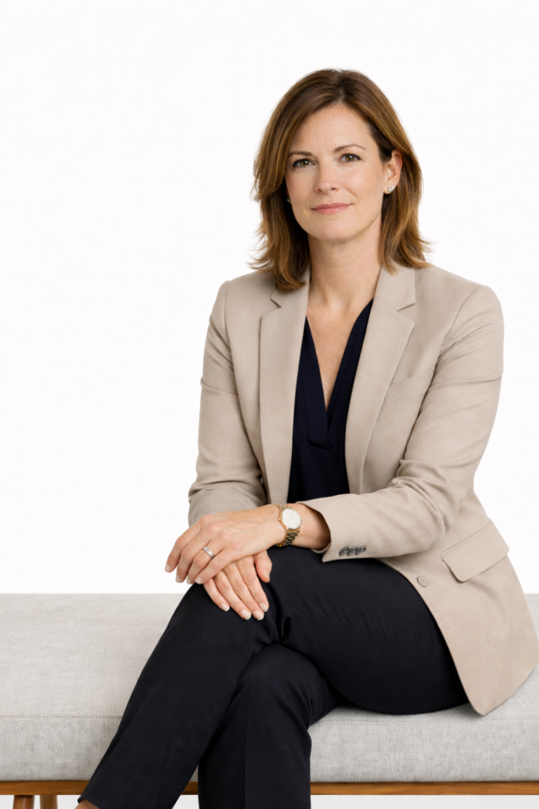 Executive woman in a beige blazer and dark trousers sits on a bench against a white background.